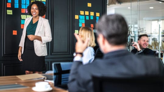 A woman stands smiling by a wall of sticky notes, leading an Information Management and Governance FAQ session.