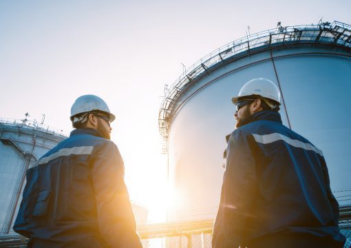 Two workers wearing hard hats and safety jackets stand outside near large industrial storage tanks at sunset, inspecting the facility as part of change management efforts.