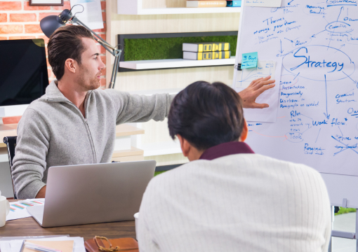 Two people in a modern office discuss an information strategy plan. One gestures toward a whiteboard covered in handwritten notes and diagrams, while the other listens. A laptop and office supplies are visible on the table.