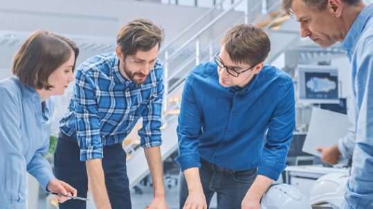 Four people stand around a table in a modern office, collaborating and looking at documents or plans, with white helmets visible on the table.