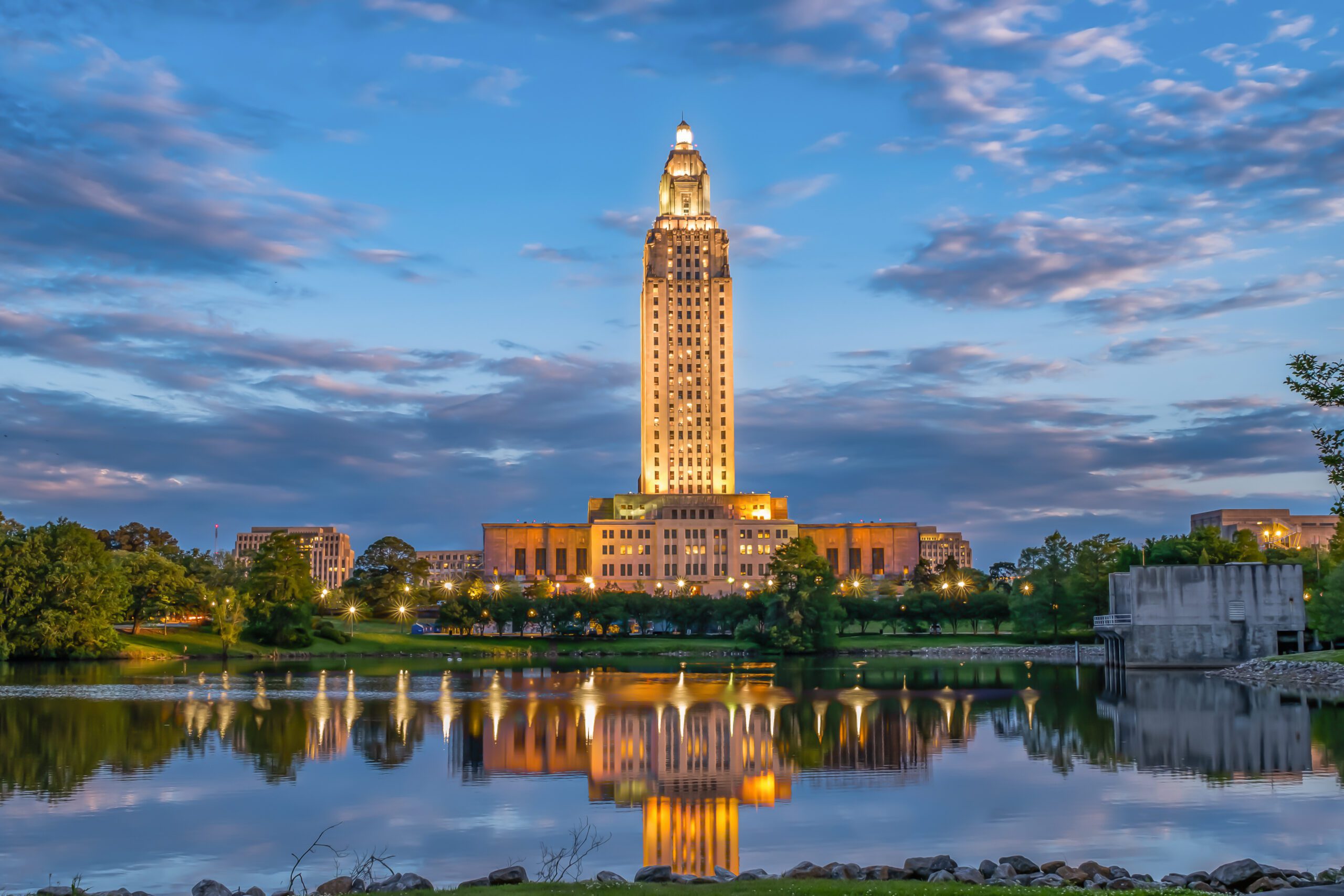A tall, illuminated building stands by a lake at dusk, reflecting in the water. Green trees and a partly cloudy sky surround the scene, creating a peaceful cityscape.